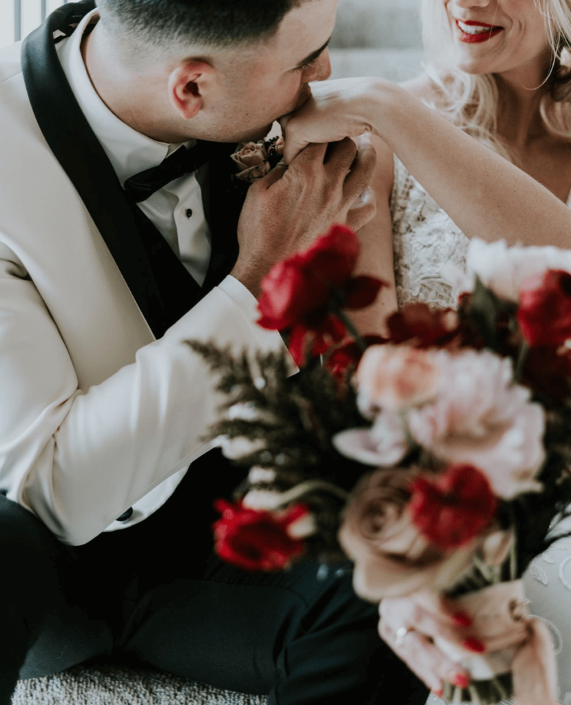 Groom kissing bride's hand and holding bouquet of red roses